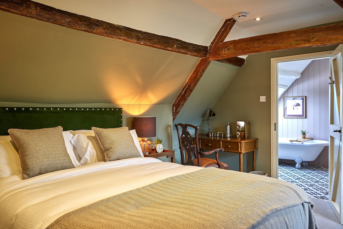 A bedroom with a large bed, wooden beams, a nightstand with a lamp, and a view into the bathroom.
