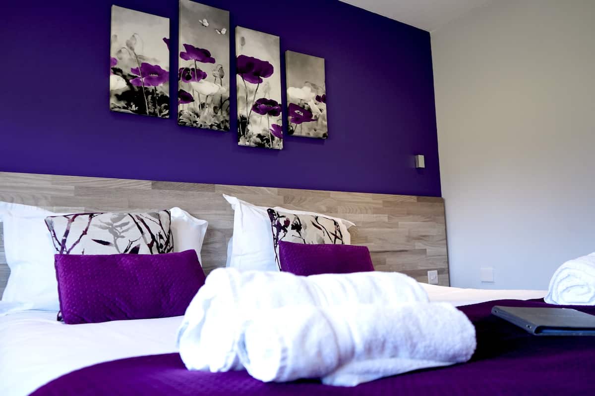 Un lit bien fait, avec une literie violette et des serviettes blanches, une tête de lit en bois et trois tableaux représentant des motifs floraux violets et blancs accrochés au mur.