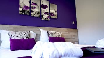 A neatly made bed with purple bedding and white towels, a wooden headboard, and three purple and white floral paintings on the wall.