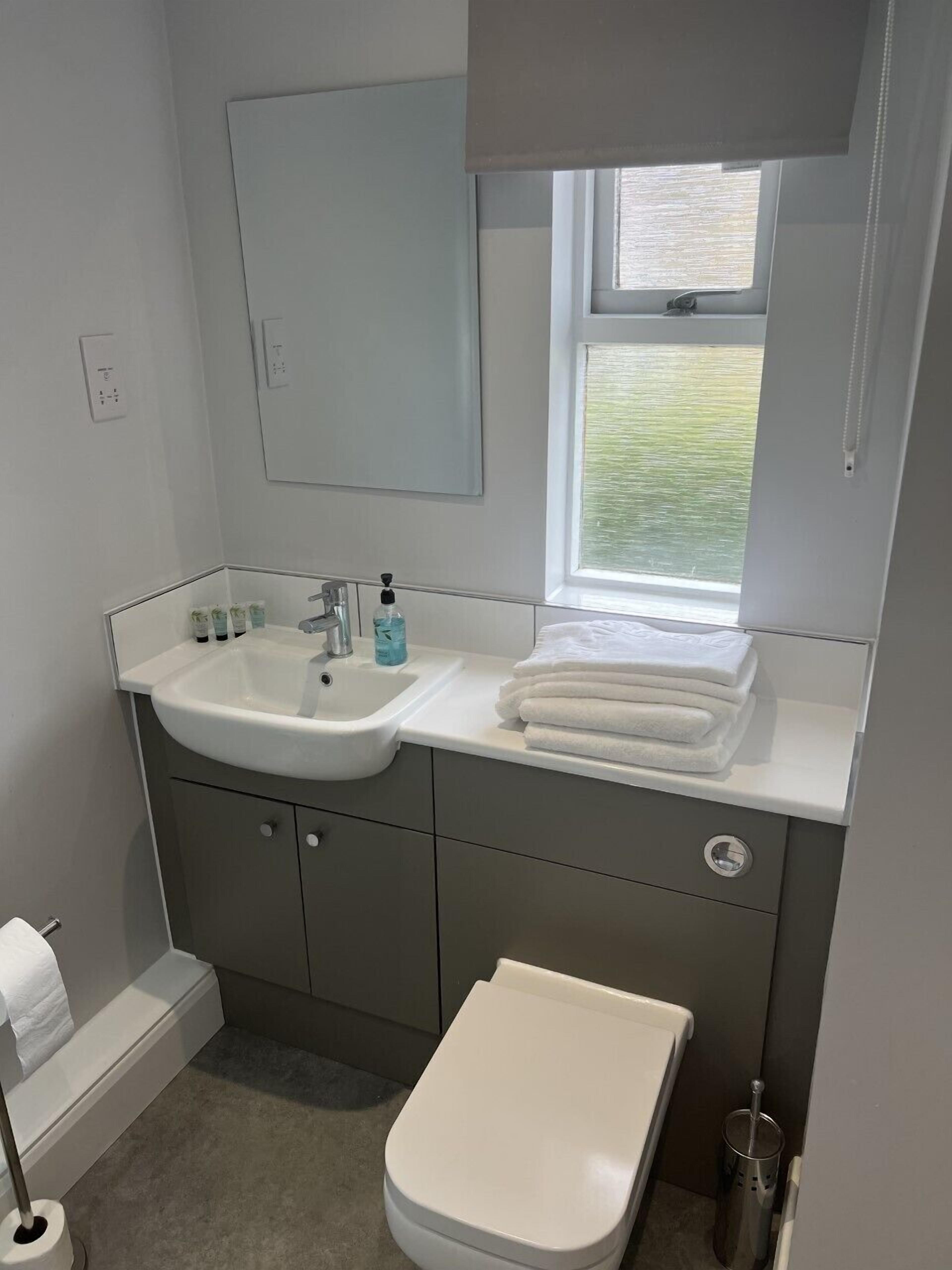 A bathroom with a white sink, mirror, and toilet. There is a window above the sink, and towels are stacked on the counter.
