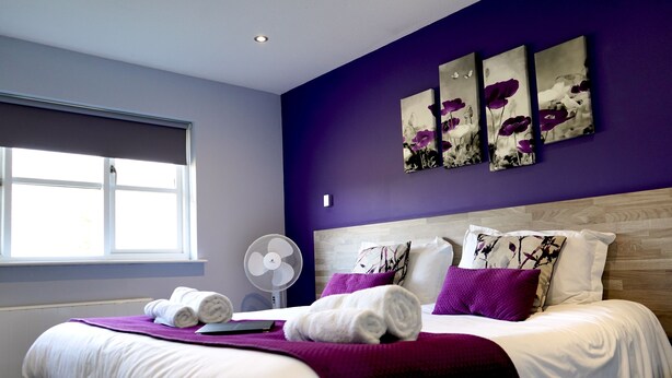 Een net opgemaakt bed met witte lakens en paarse accenten, een raam met zonwering en een ventilator bevestigd aan de muur.
