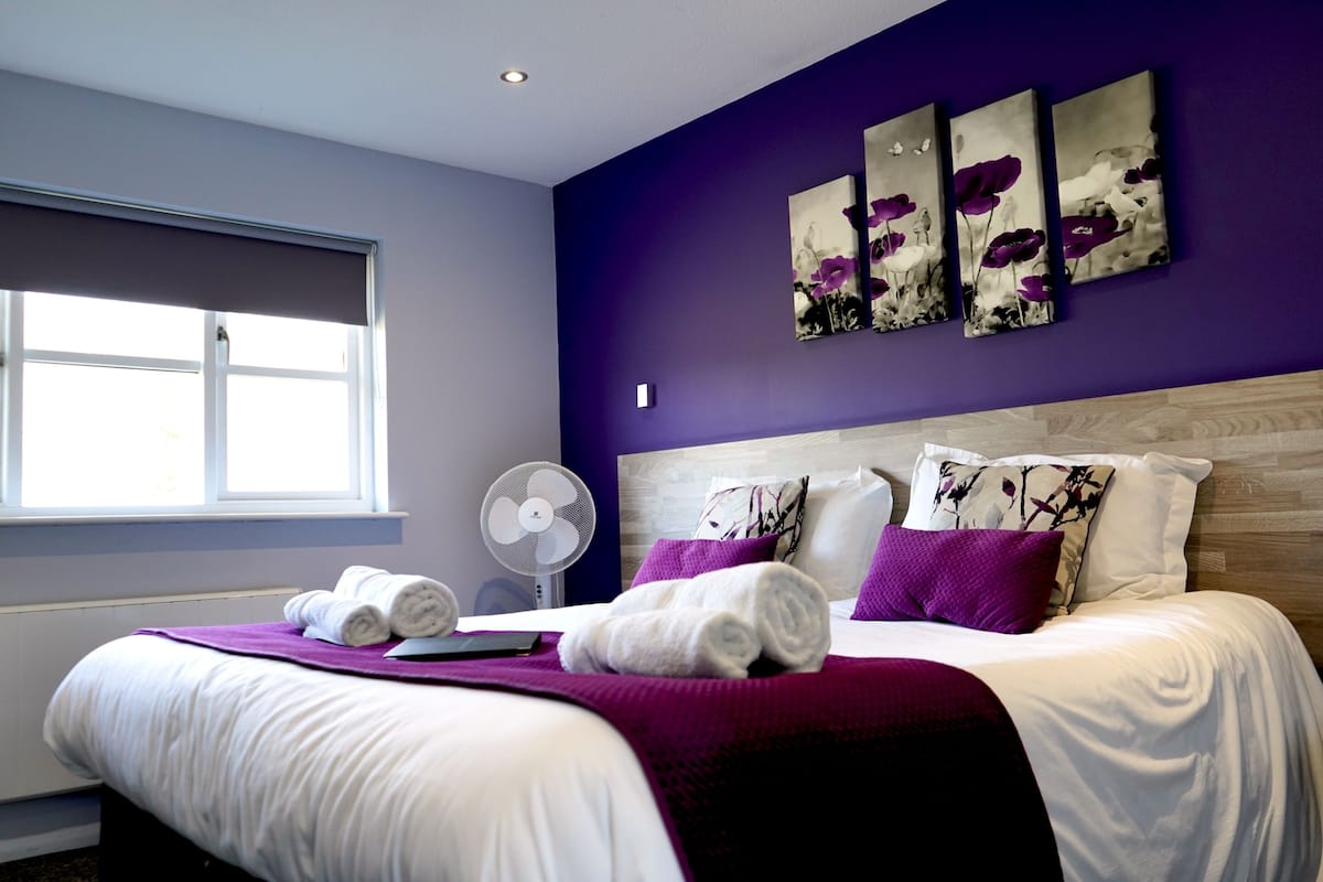 Un lit bien fait, avec du linge de lit blanc et des touches de couleur violette, une fenêtre avec des stores et un ventilateur fixé au mur.