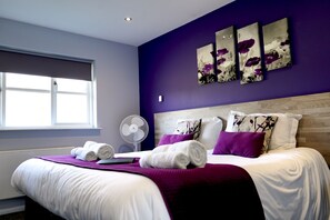 A neatly made bed with white linens and purple accents, a window with blinds, and a wall-mounted fan. - The Woolpack Inn Islip (Kettering)