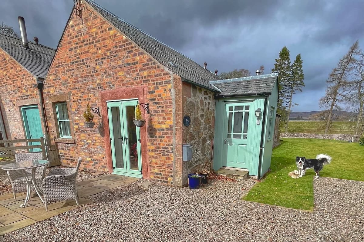 A brick house with a green door, a small attached shed, and a dog in the yard.