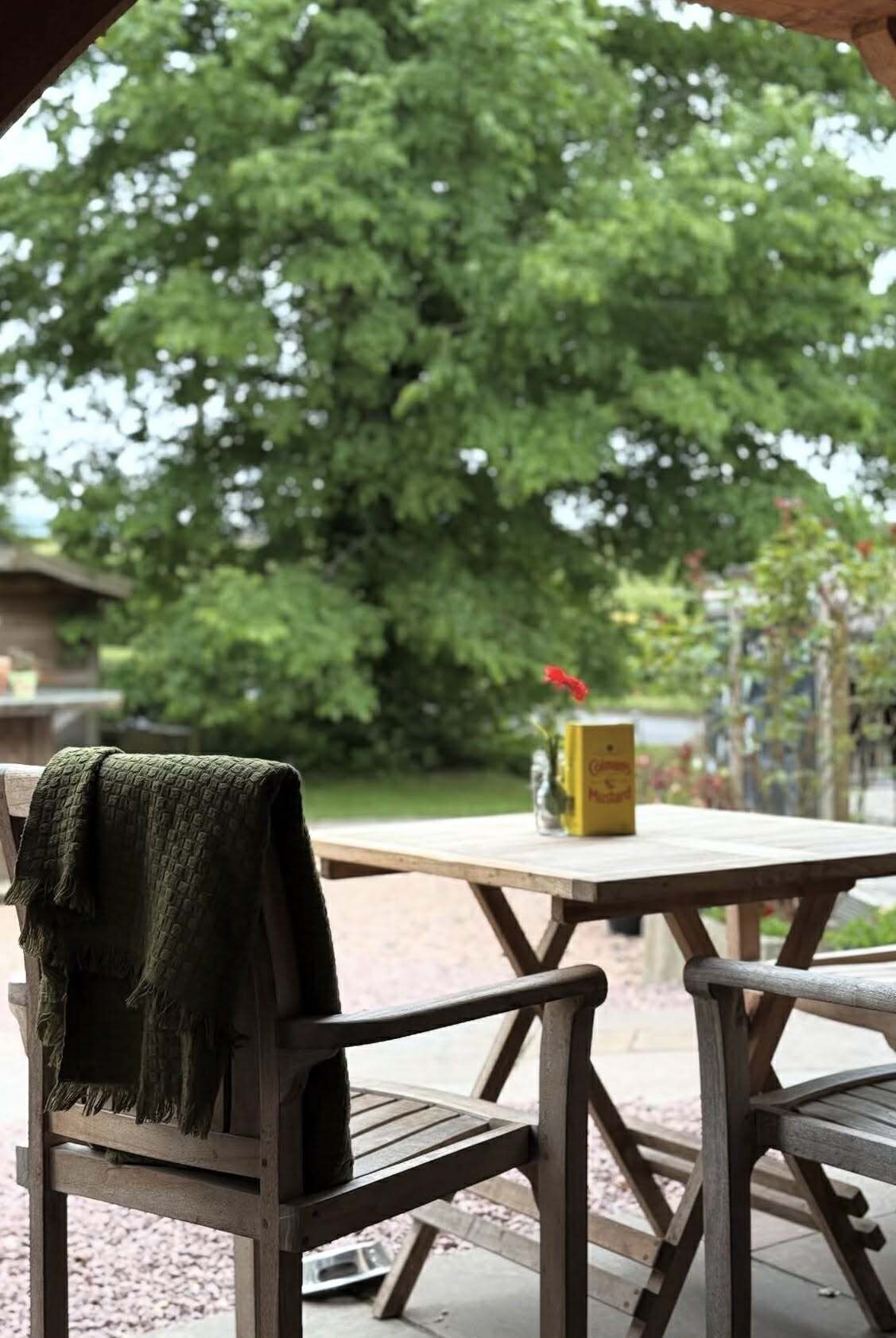 A wooden bench with a green blanket, a wooden table with a yellow box and a red flower vase, and a large tree in the background.