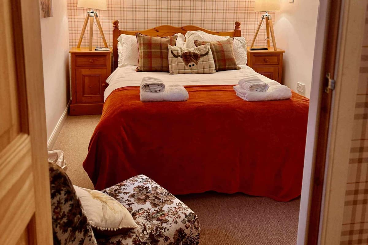 A bedroom with a red bedspread, a wooden headboard, two bedside lamps, a plaid-patterned wall, and a small chair with a floral cushion.