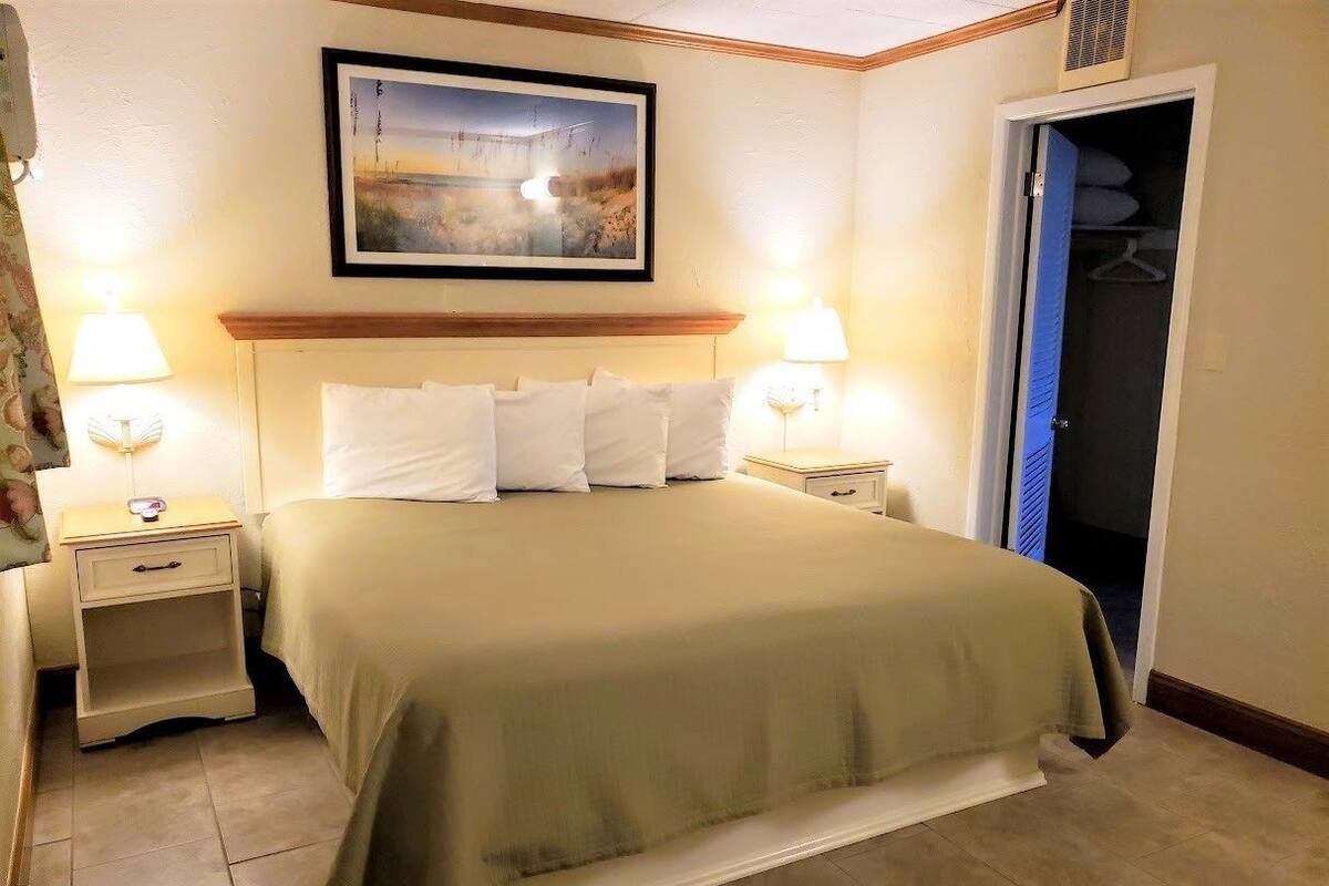 Une chambre d’hôtel avec un lit, deux tables de chevet, une commode, une lampe, un tableau encadré au mur et un placard avec une porte bleue.