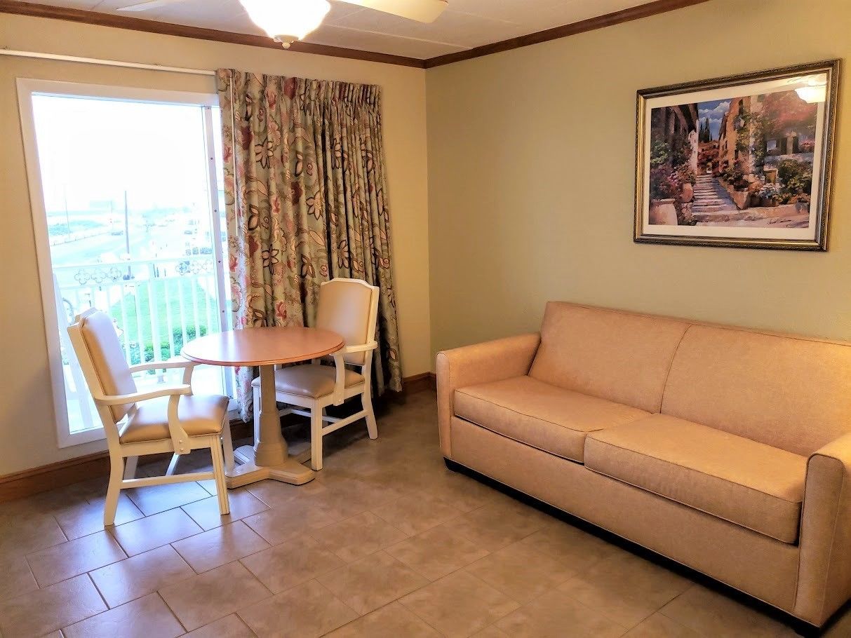 A living room with a beige sofa, a wooden dining table with two chairs, and a large window with floral curtains.