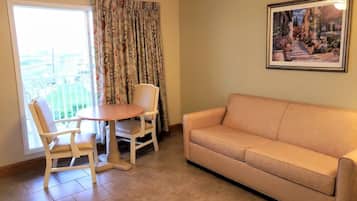 A living room with a beige sofa, a wooden dining table with two chairs, and a large window with floral curtains.