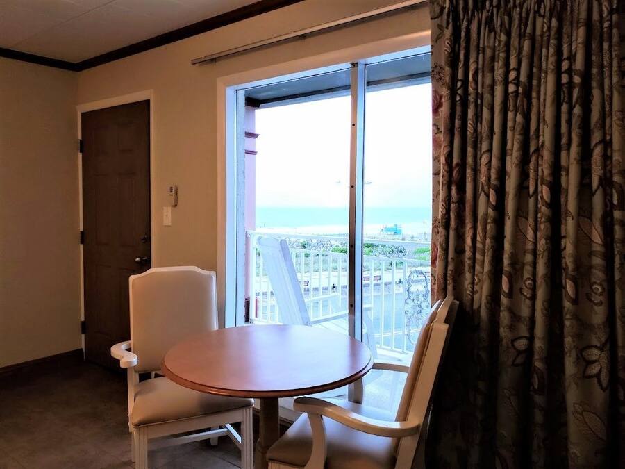 Quarto com uma cadeira branca e uma pequena mesa redonda, uma porta e vista através de uma janela grande com cortinas.
