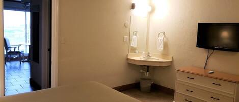 A hotel room with a bed, a sink, a mirror, a television, and a view of the outside balcony.