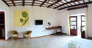 Una habitación con una cama, dos sillones de mimbre, un escritorio con silla, un televisor montado en la pared y un ventanal con cortinas.
