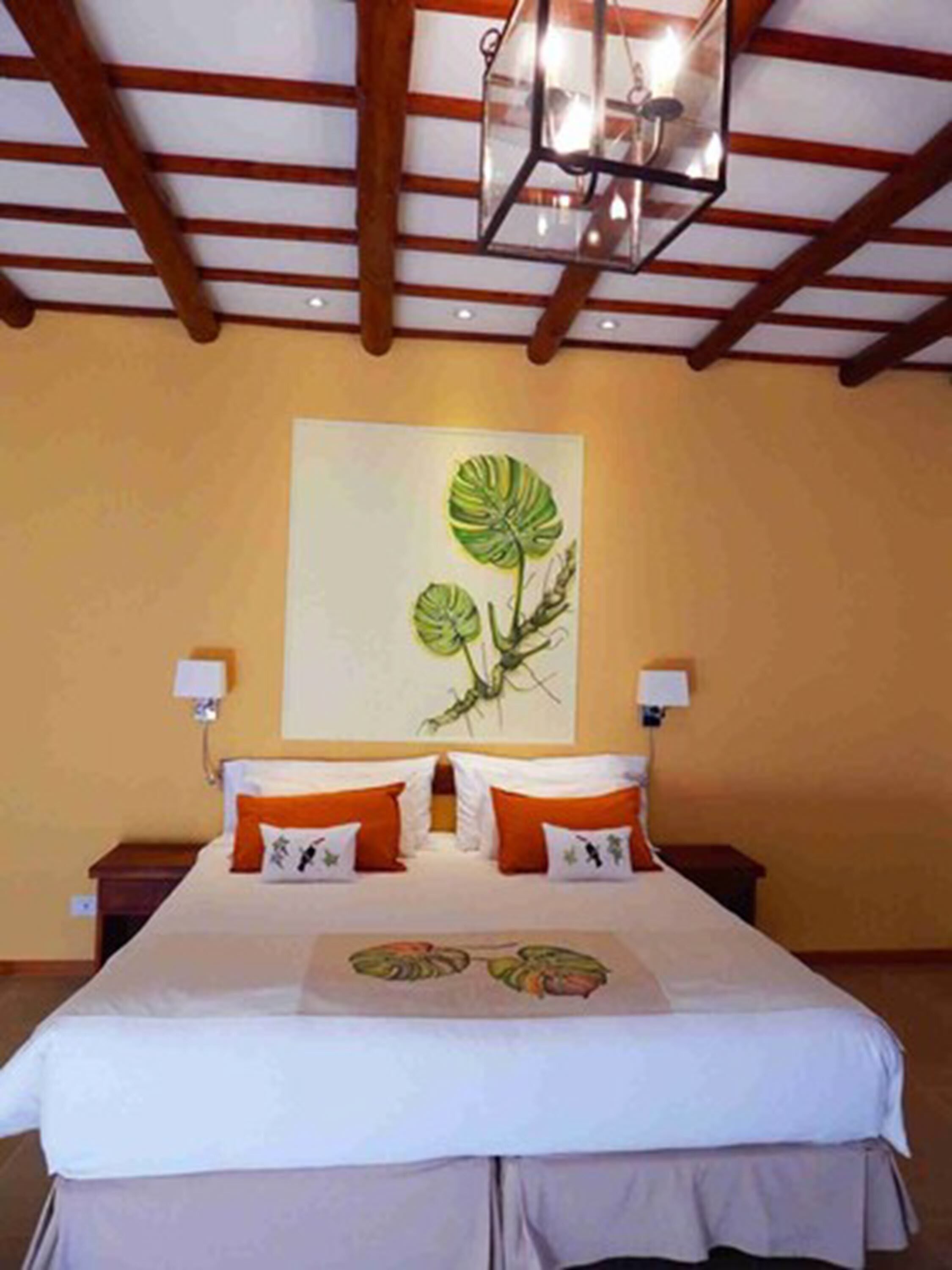 Habitación de hotel con cama, dos mesitas de noche, una lámpara de pared y una ilustración de planta grande en la pared.