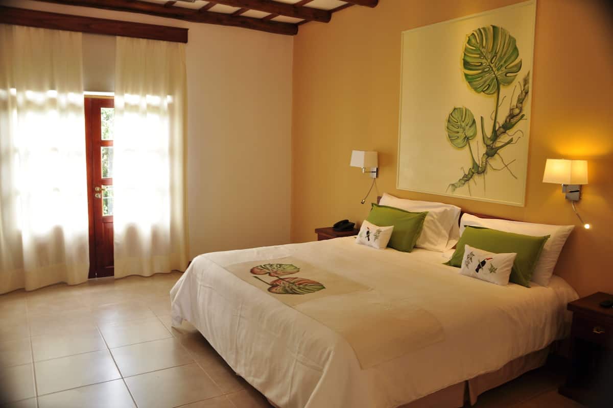A hotel room with a bed, a nightstand, a lamp, a painting on the wall, and a window with curtains.