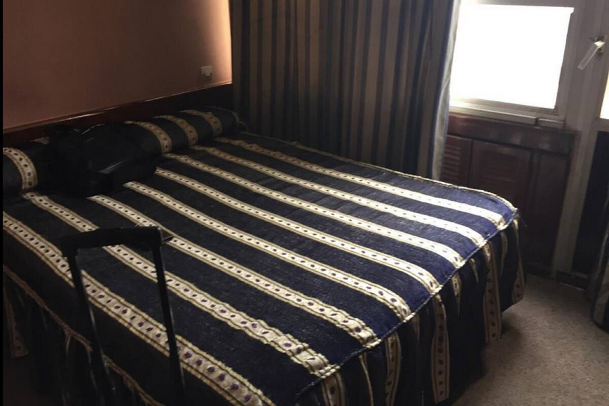 A small, single-bed room with a striped bedspread, a wall-mounted light, and a window with curtains.