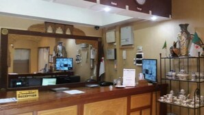 A reception area with a wooden counter, two computer monitors, a flag, and a decorative shelf with items. - Hotel Roza (Algiers)