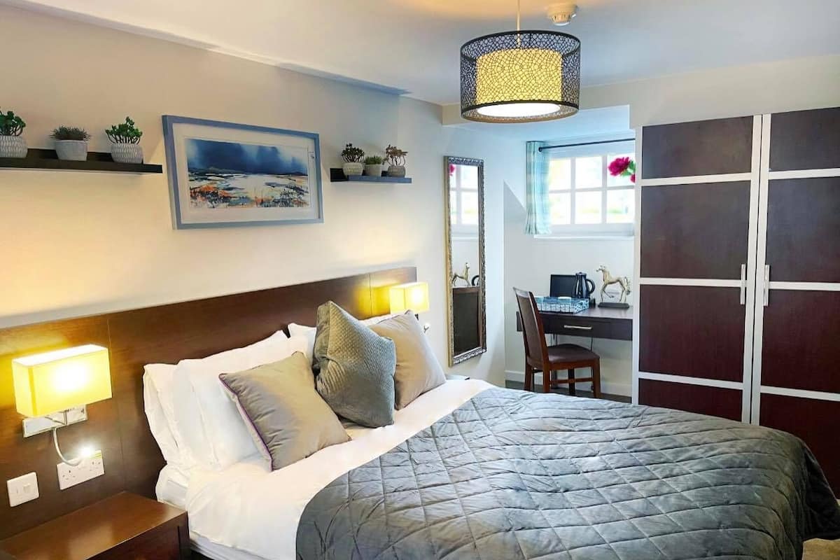 A hotel room with a bed, bedside lamps, a desk, a chair, a wardrobe, and a large window.