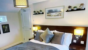 A neatly made bed with a quilted grey duvet, two bedside lamps, a framed picture on the wall, and a shelf with potted plants. - The High Flyer (Ely)