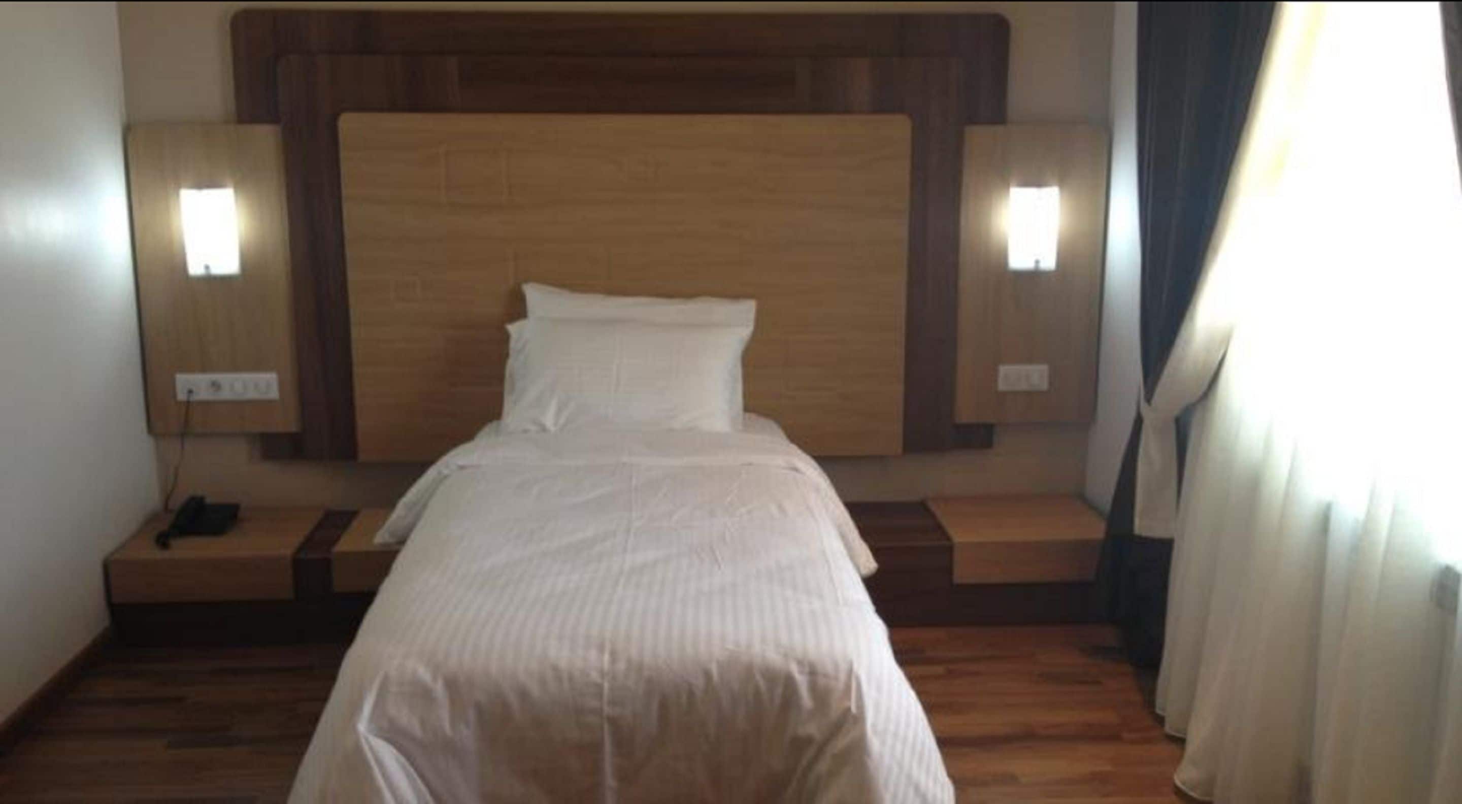 Une chambre d’hôtel avec une tête de lit en bois, un lit recouvert de draps blancs, deux tables de chevet et des lampes fixées au mur.