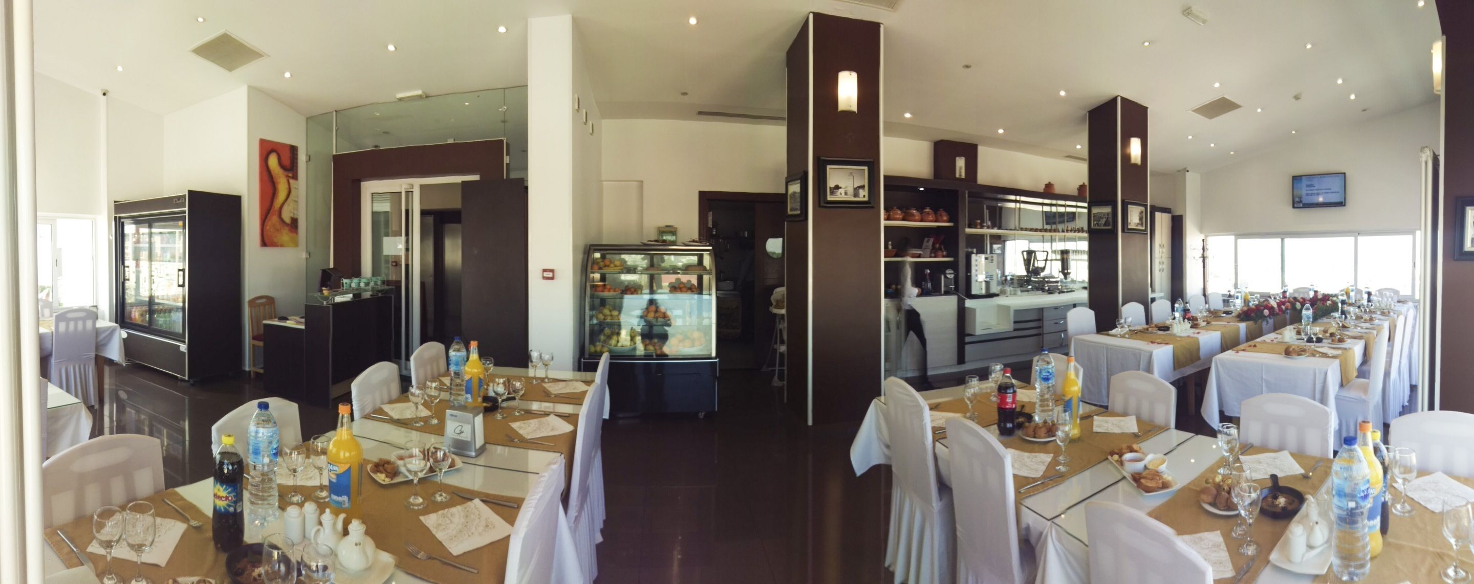 Un comedor de hotel con varias mesas dispuestas para los huéspedes, una zona de cocina y un mostrador con productos alimenticios.