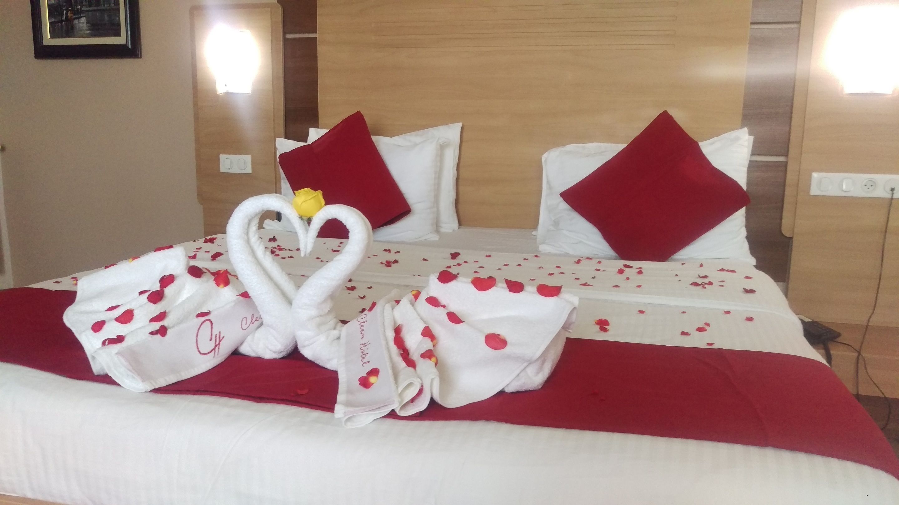Una camera d'albergo con un letto rifatto, lenzuola bianche, cuscini rossi e un'accorto allestimento di asciugamani a forma di cuore con una rosa.