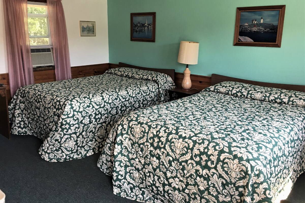 Une chambre avec deux lits, une fenêtre avec des rideaux, une lampe et deux tableaux encadrés au mur.