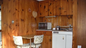 Cocina compacta con paredes de madera, microondas, lavamanos y un refrigerador pequeño. Hay dos sillas tapizadas y una mesa.