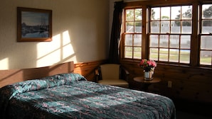 Una habitación con una cama, una silla, una mesita con un florero y un ventanal con vistas al exterior.
