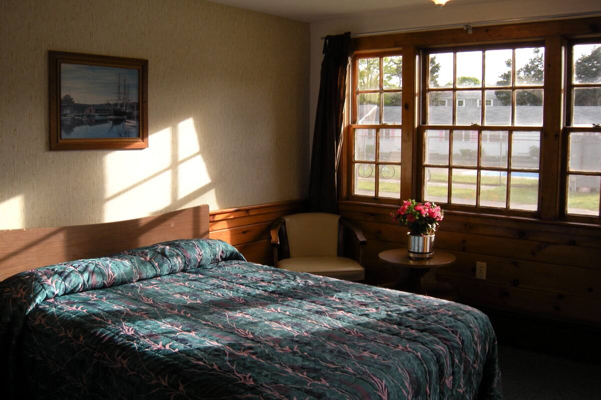 Une pièce comprenant un lit, une chaise, une petite table avec un vase de fleurs et une fenêtre donnant sur l’extérieur.