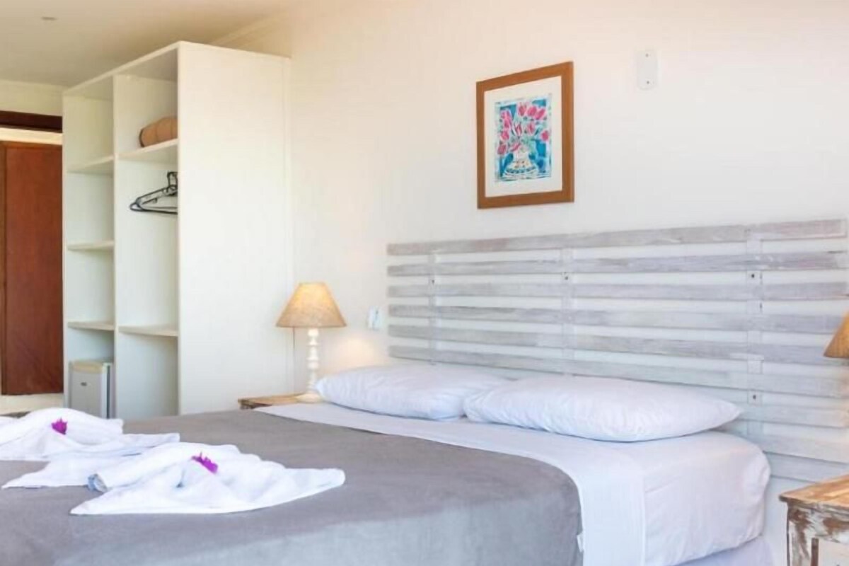 A neatly made bed with a grey bedspread and white linens, a bedside table with a lamp, and a wall-mounted shelf with a framed picture.