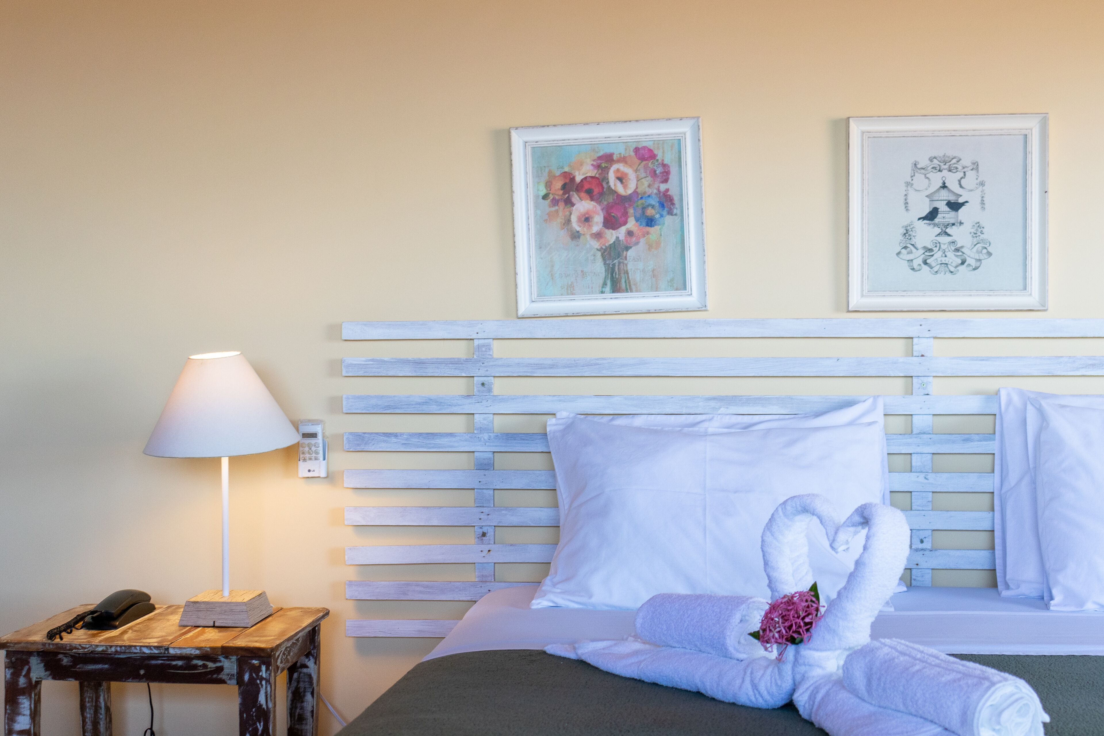A hotel room with a wooden headboard, a bedside table with a lamp, a phone, and a bed with white linens and a heart-shaped towel arrangement.