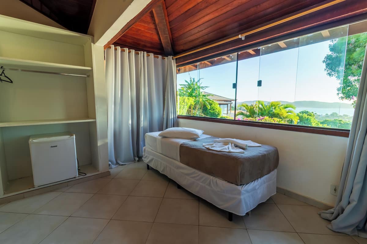 Ein Schlafzimmer mit einem Bett, einem kleinen weißen Gerät, einem Fenster mit Vorhängen und Blick ins Grüne.