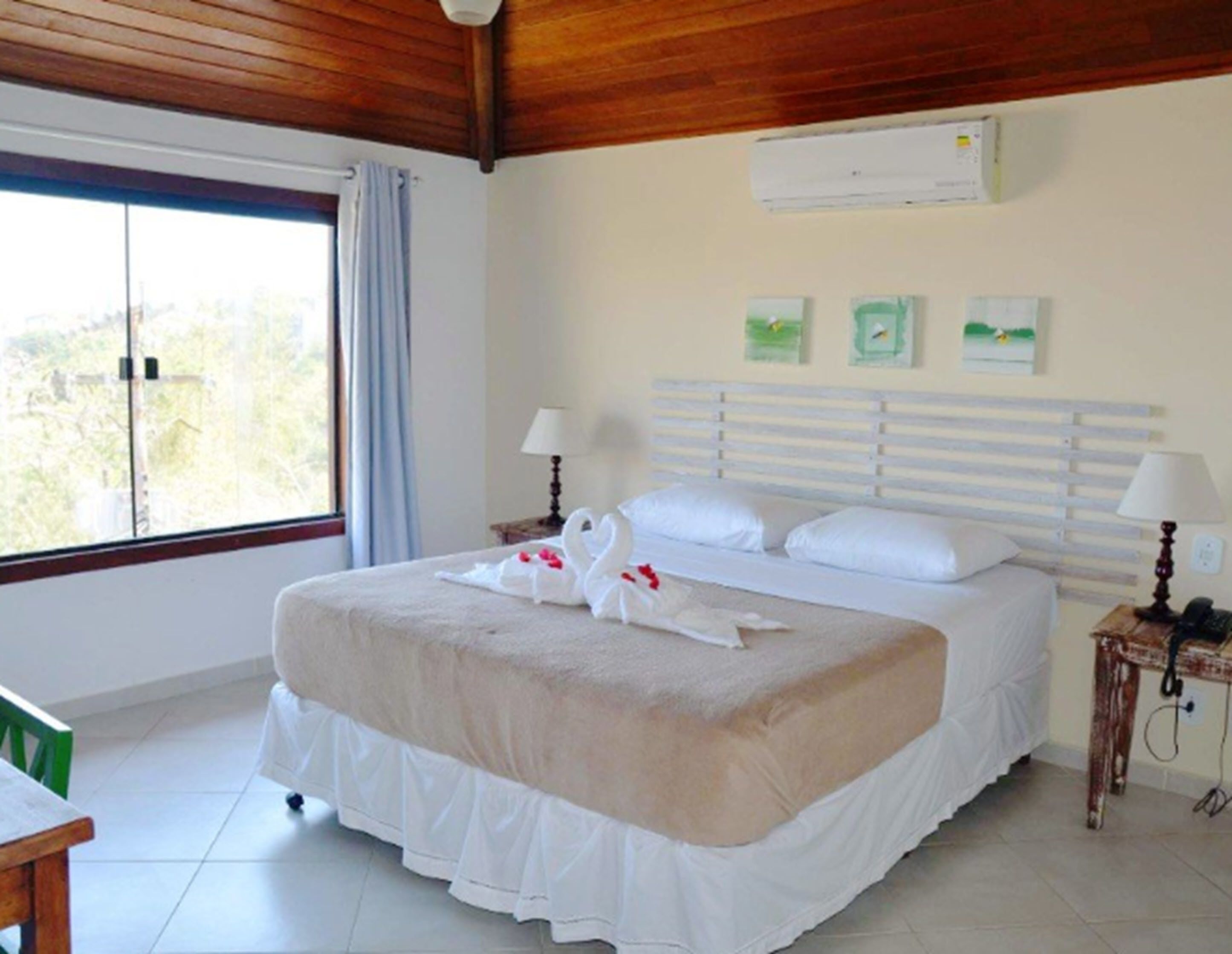 A hotel room with a large bed, a wooden ceiling, a window with a view, a small table, and a nightstand with a lamp.