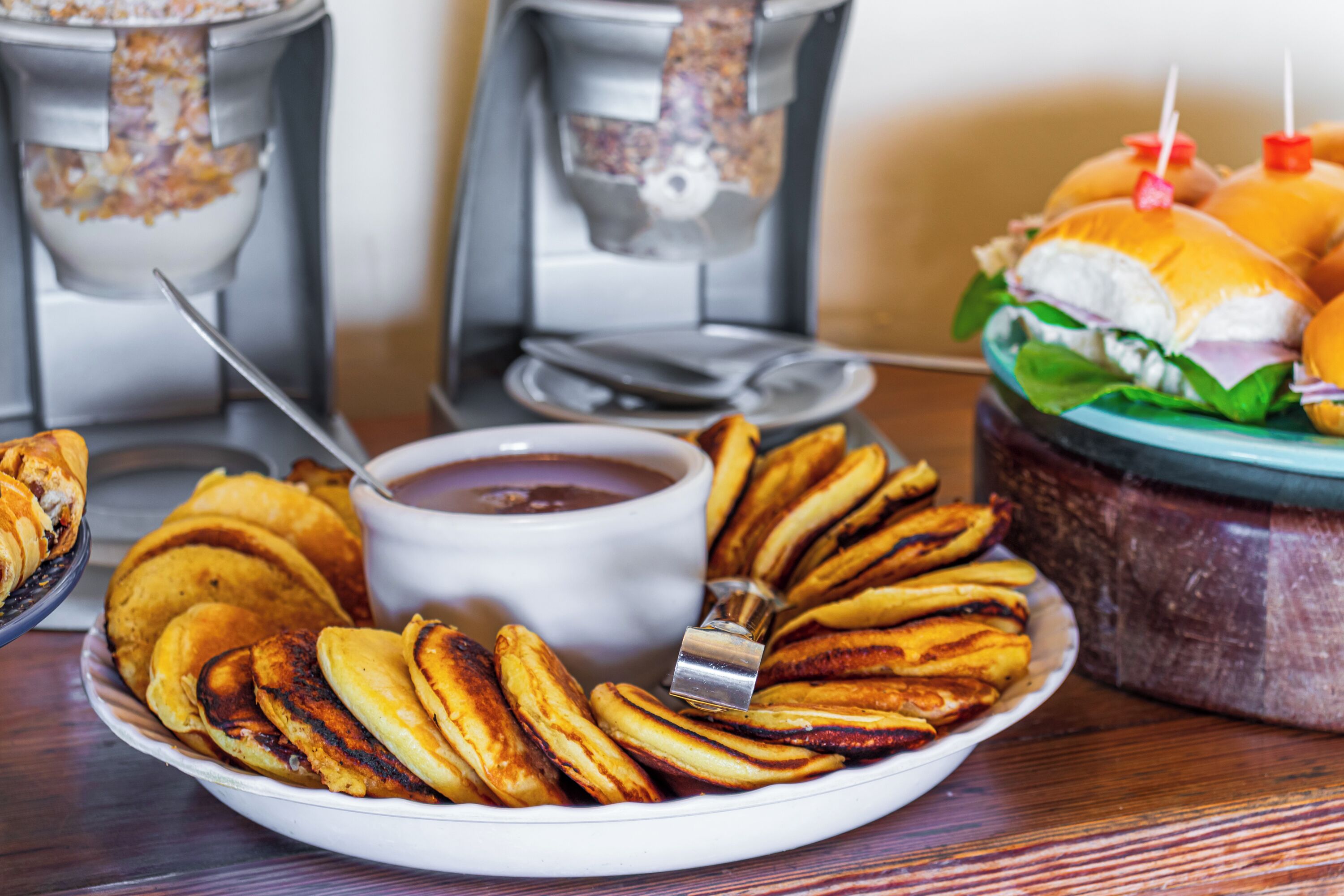 A plate of fried plantains, a bowl of dipping sauce, and a basket of sandwiches on a wooden table.