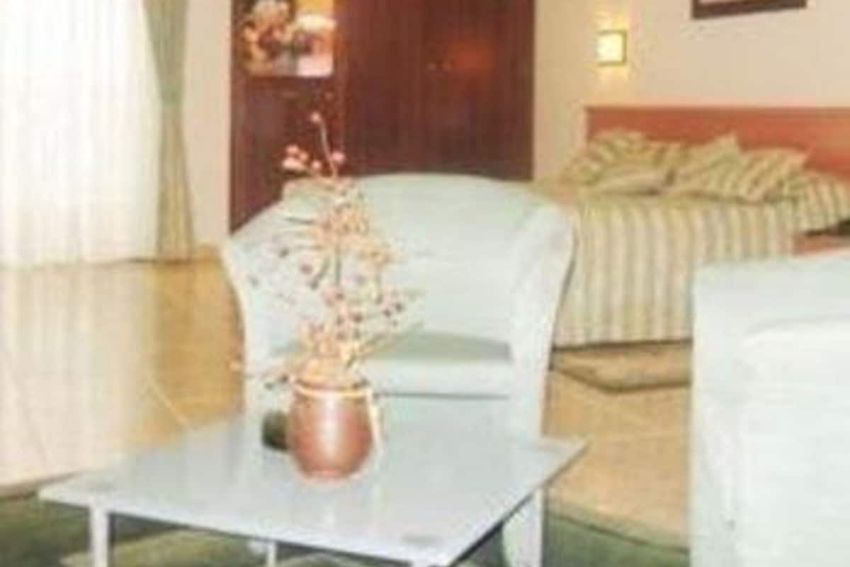 A hotel room with a bed, a chair, a table with a vase, a painting on the wall, and a door.