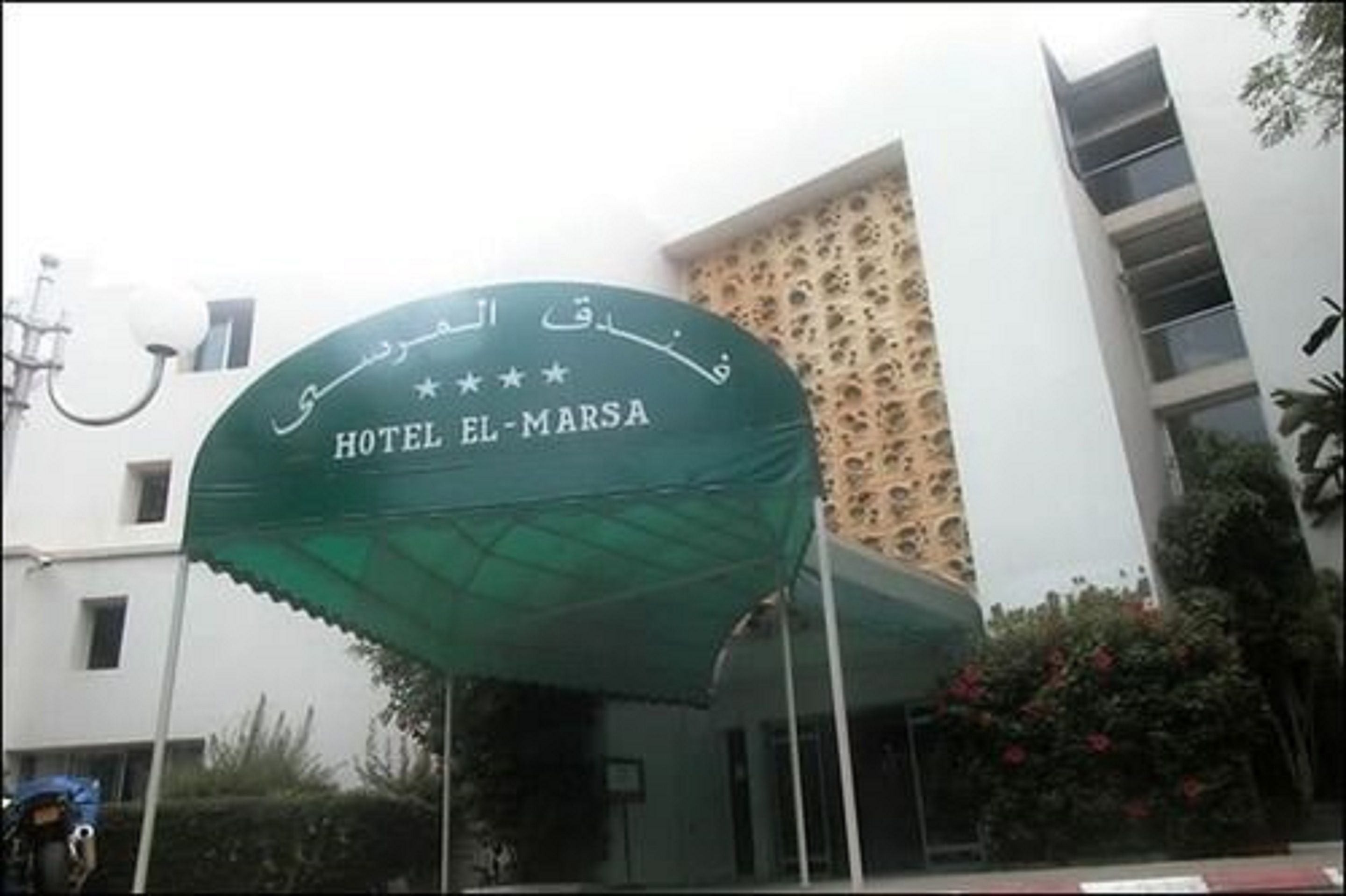 Entrada de un hotel con un toldo verde que muestra el nombre 'HOTEL EL-MARSA' en árabe e inglés.