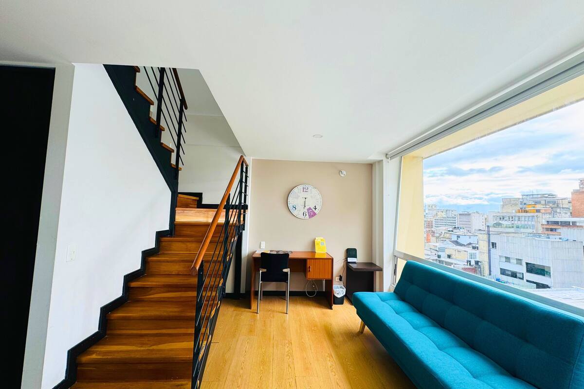 A modern apartment with a wooden staircase, a blue sofa, a desk with a chair, and a city view through large windows.