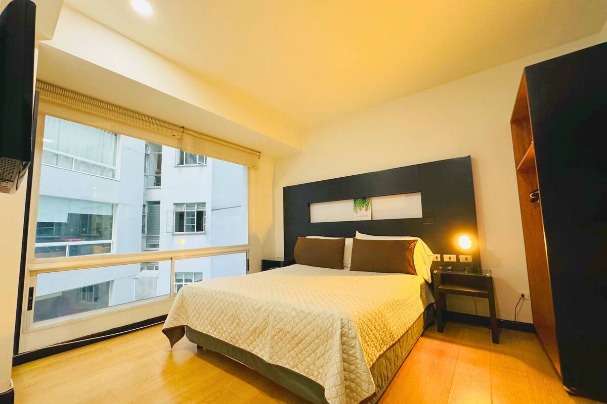 A hotel room with a large bed, a nightstand, a wall-mounted shelf with a plant, and a view of the building outside.