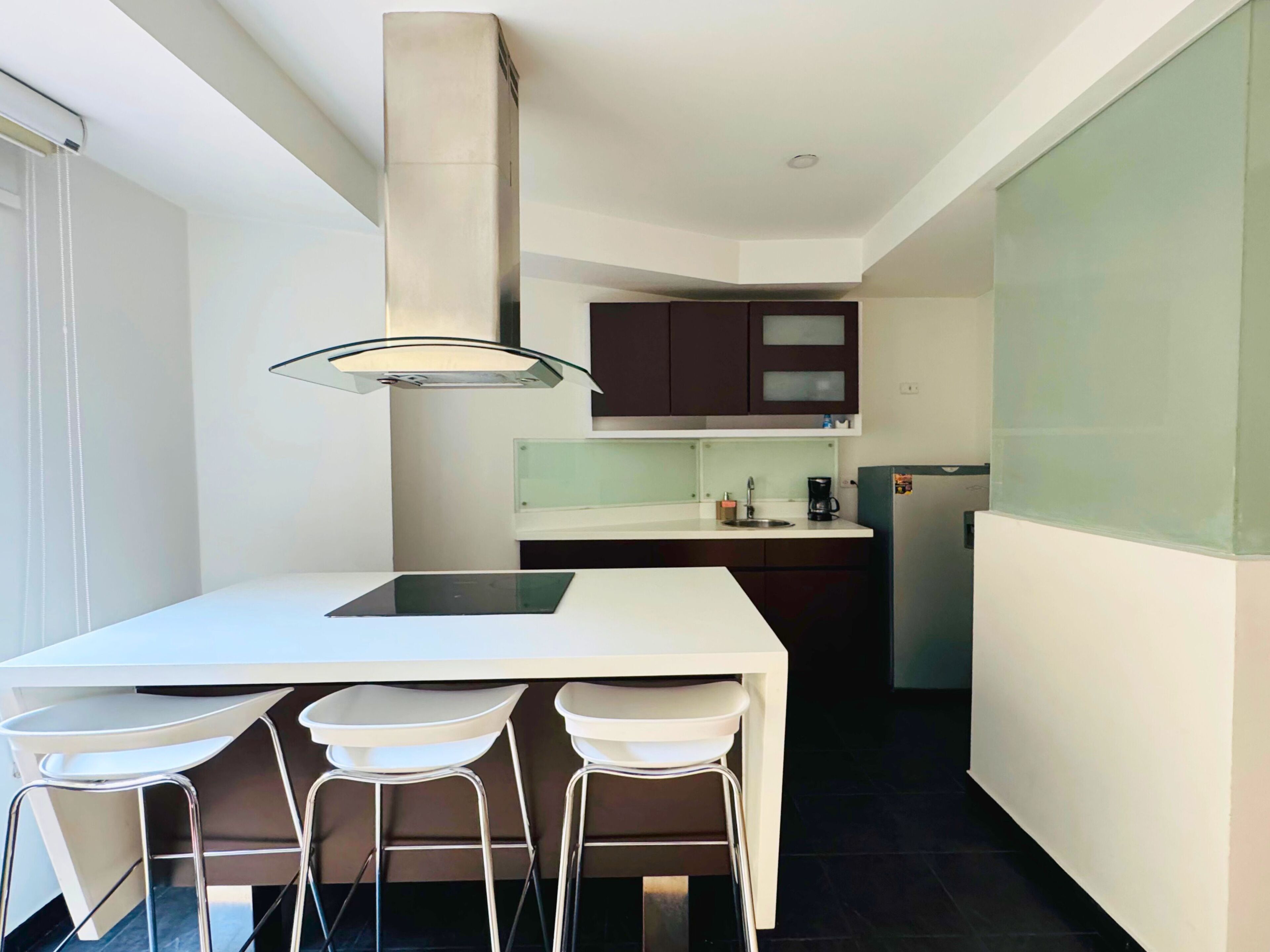 A modern kitchen with a white countertop, dark cabinets, and a stainless steel refrigerator. There are three white bar stools at the kitchen island.