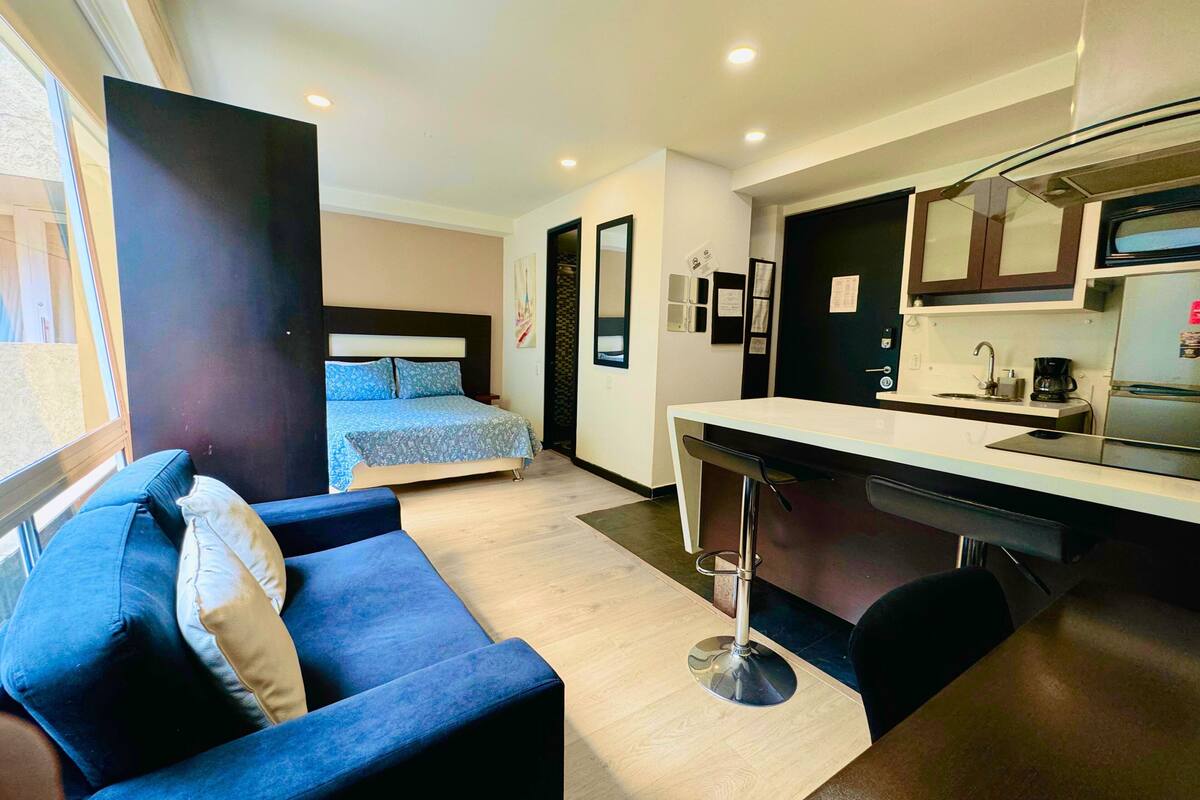 A modern hotel room with a bed, a blue sofa, a kitchenette, and a view of the outside.