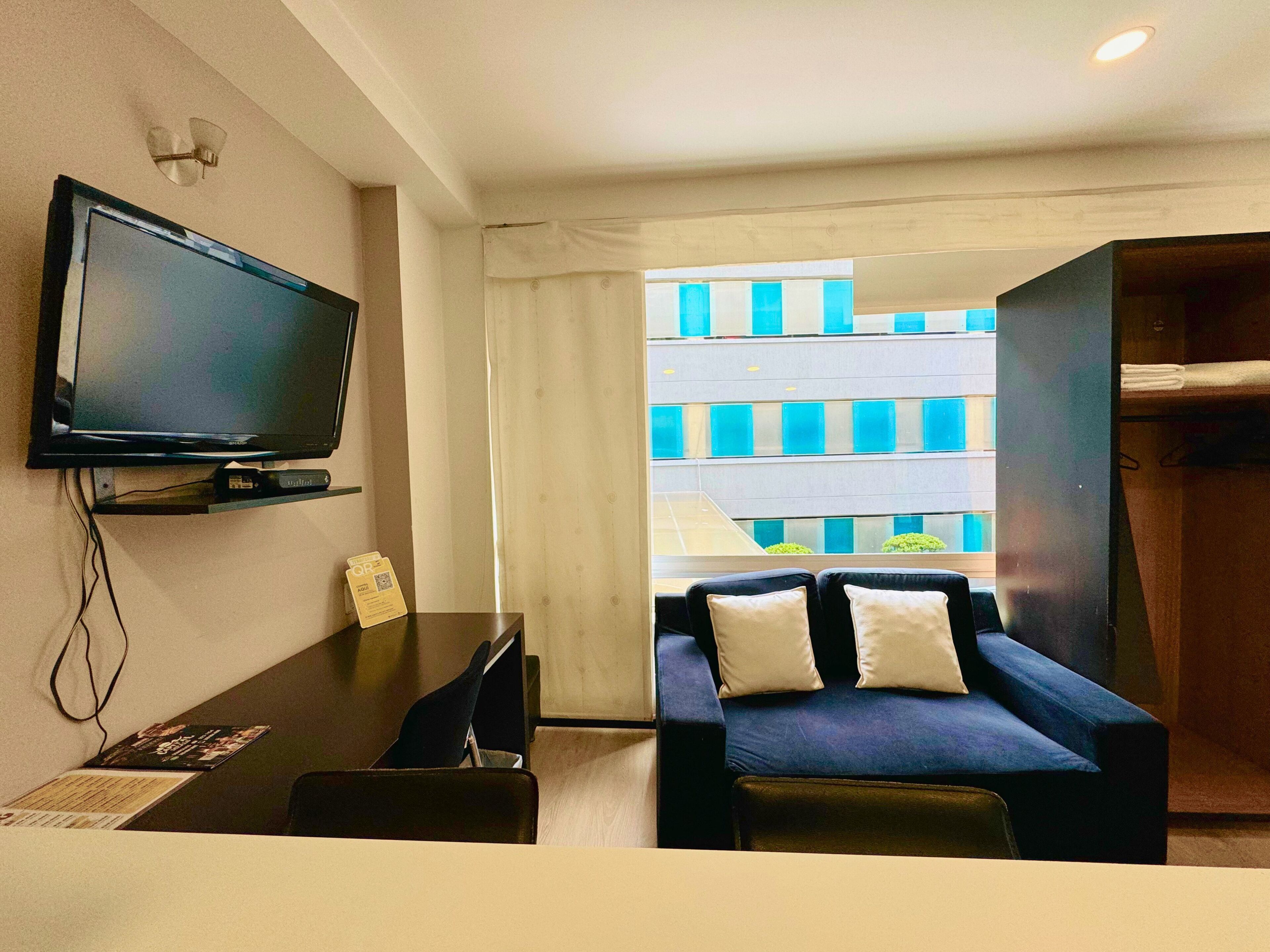A modern hotel room with a flat-screen TV mounted on the wall, a dark blue sofa with two pillows, a small desk with a chair, and a view of a building with blue and white windows.