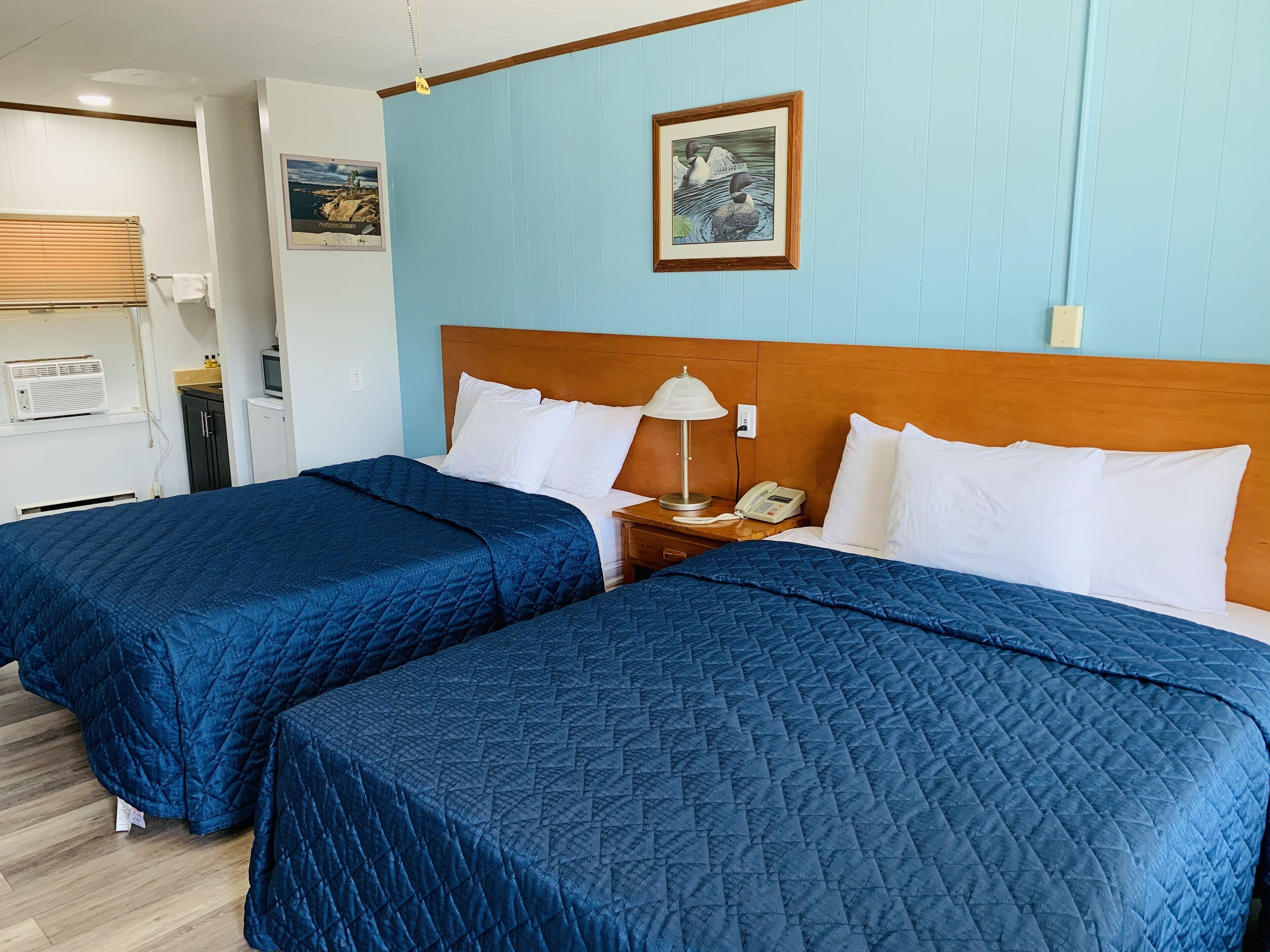 A hotel room with two beds, a wooden headboard, a bedside table with a lamp, a phone, and a framed picture on the wall.