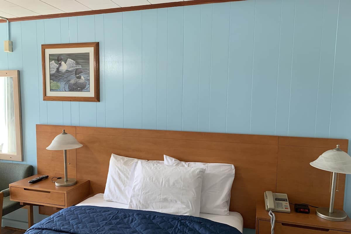 A hotel room with a wooden headboard, a blue quilted bedspread, white pillows, two bedside lamps, a framed picture on the wall, and a bedside phone.