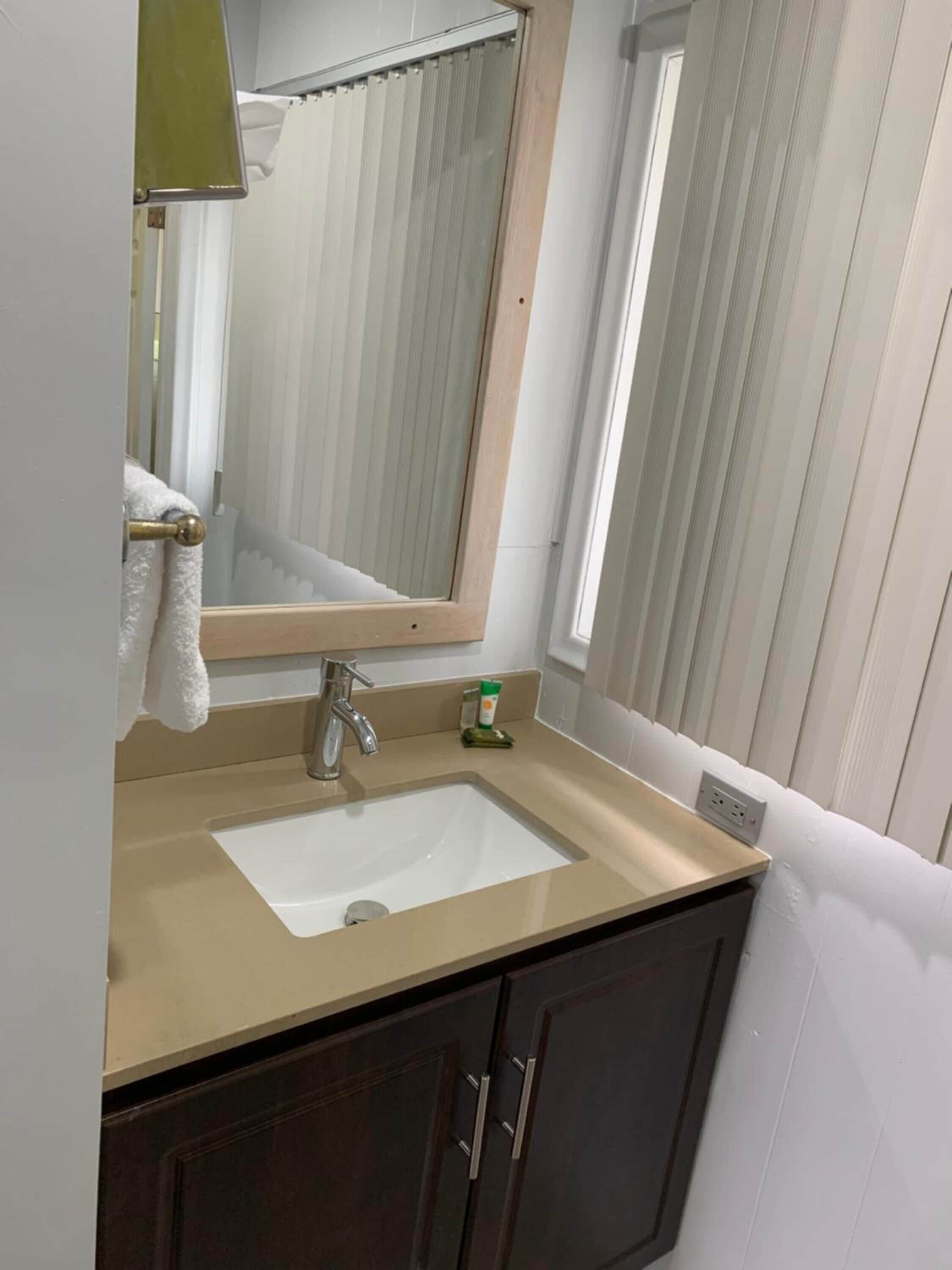 Un baño con un lavamanos color beige, armarios de madera oscura, un espejo y una ventana con cortinas.