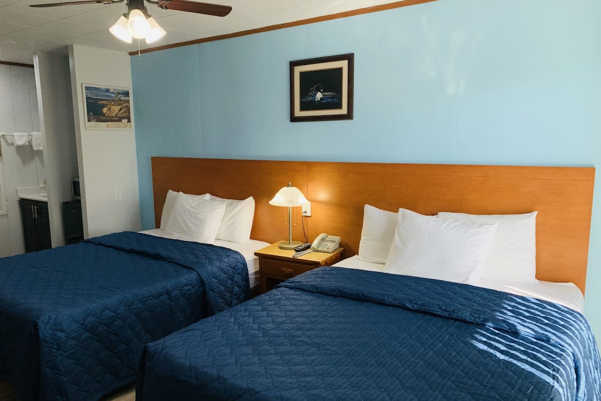 A hotel room with two beds, each with a blue quilted bedspread and white pillows. There is a wooden headboard, a bedside table with a lamp and a phone, and a framed picture on the wall.