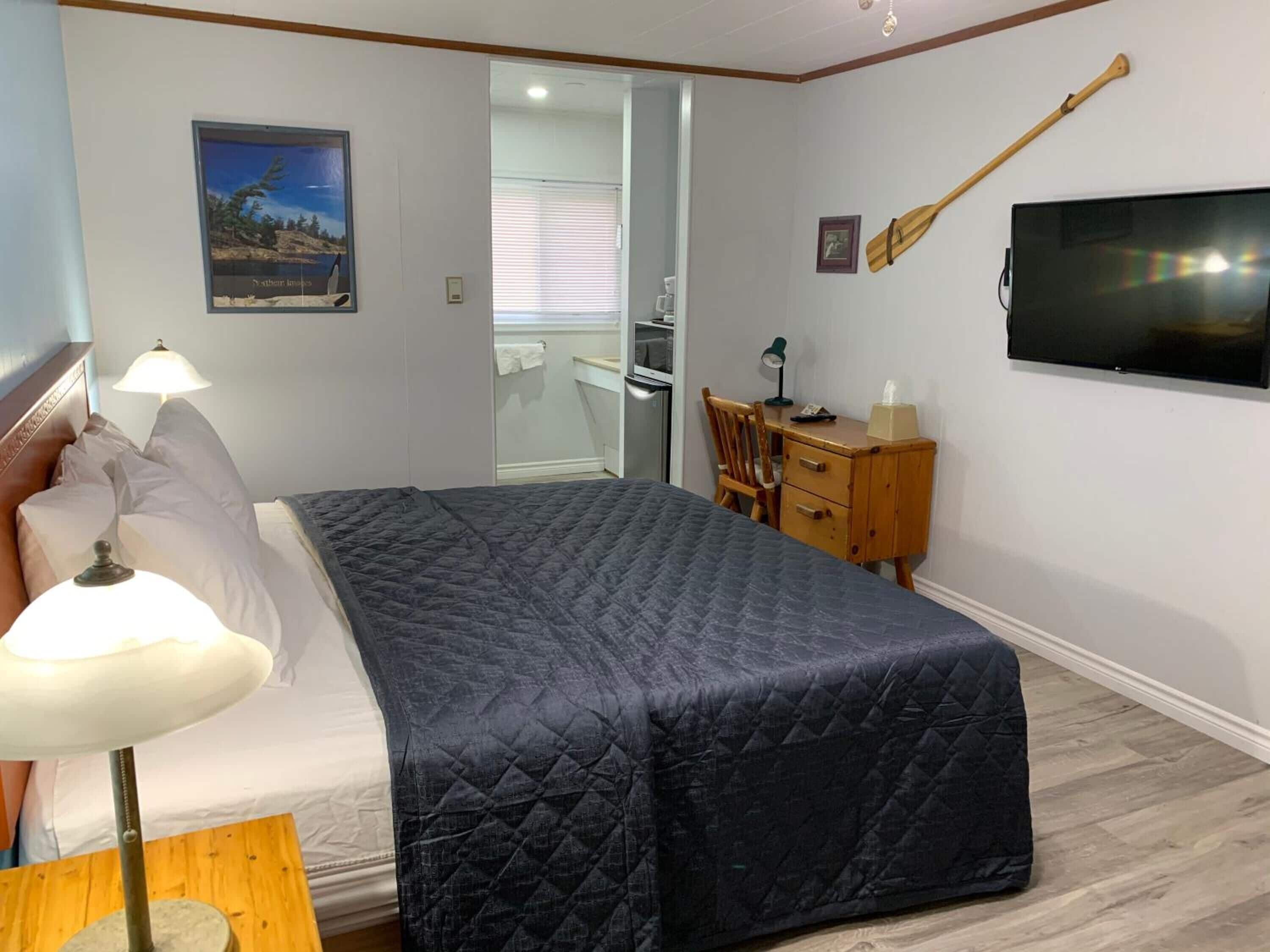 Een slaapkamer met een bed, een bedlampje, een houten bureau, een stoel, een televisie en een schilderij aan de muur.