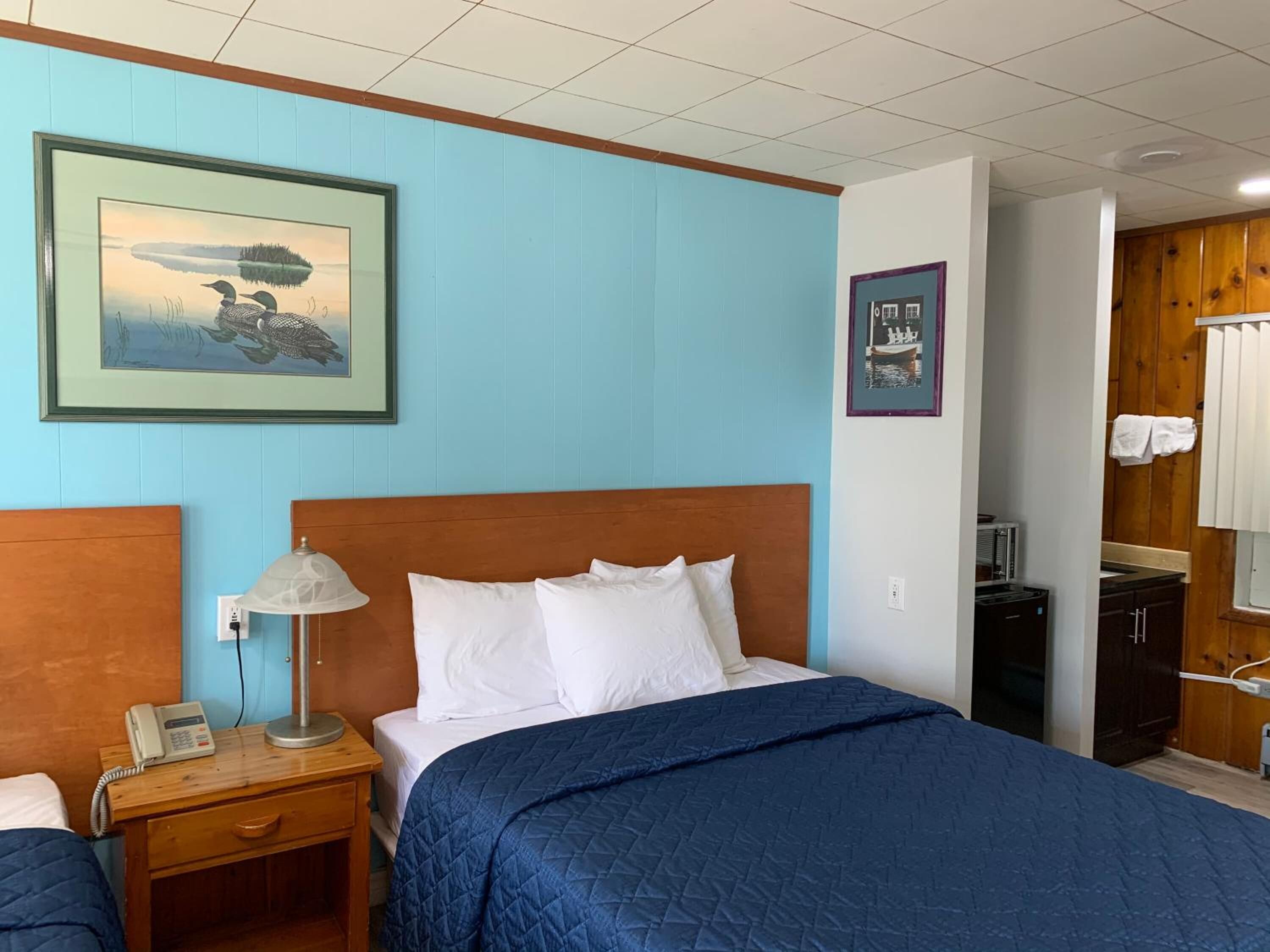 A hotel room with a wooden headboard, a blue quilted bedspread, a bedside lamp, a framed picture of a lake, and a view into the bathroom.