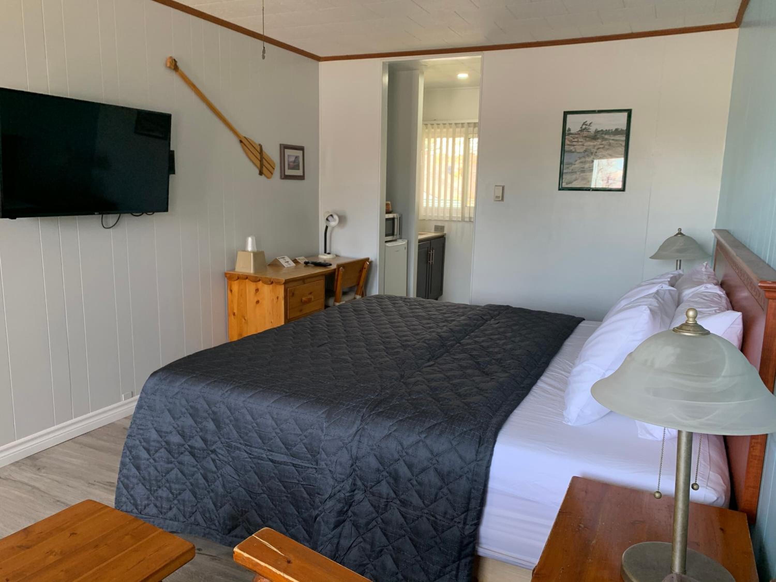 A bedroom with a bed, bedside table, lamp, desk, chair, TV, and artwork on the wall.