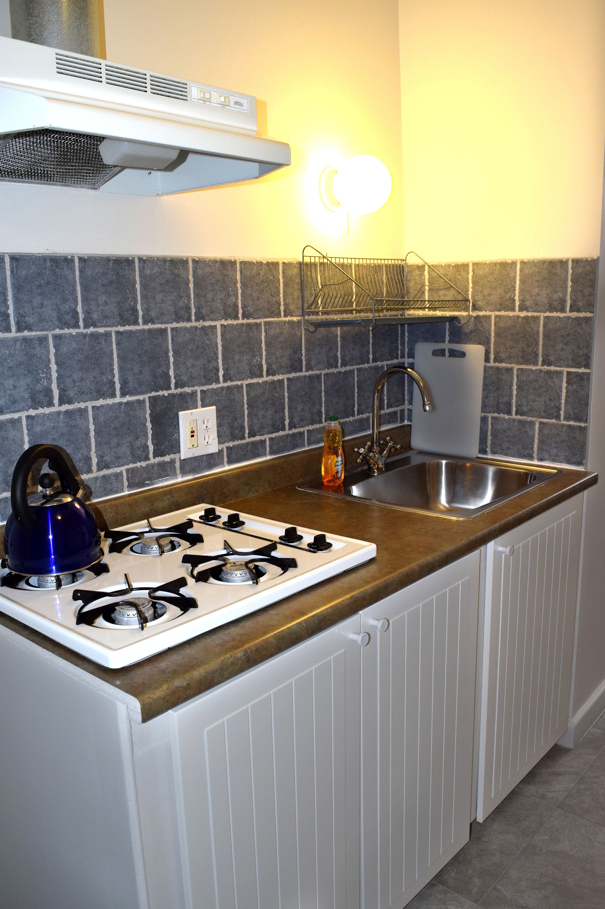 A compact kitchen with a gas stove, a kettle, a sink, and a backsplash of gray tiles.