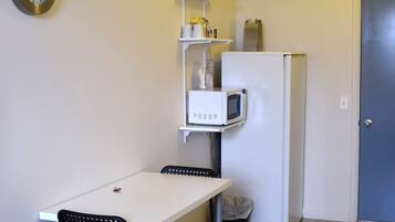 A compact kitchenette with a white refrigerator, a microwave, and a small shelf with kitchen items. There is a white table with two chairs and a clock on the wall.